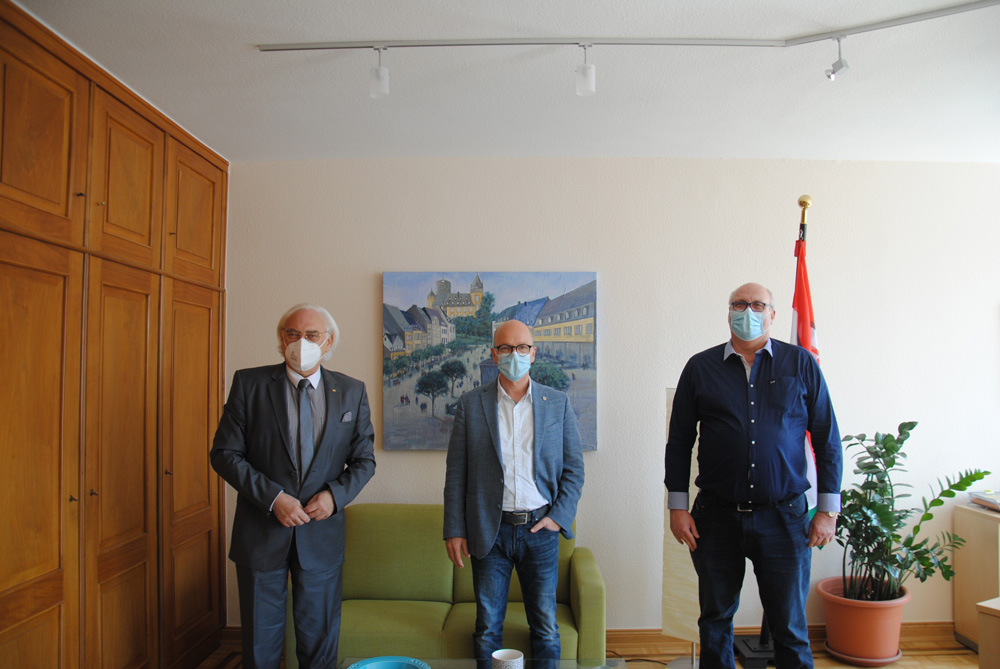 Das Bild zeigt den Hans Mayer, Dirk Meid und Hermann Wagner mit aufgesetzten Masken und Abstand in einer Reihe im Büro des Oberbürgermeisters stehend. Im Hintergrund hängt ein Gemälde der Genovevaburg.